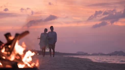 Couple with Champagne glasses walks along sandy beach by bonfire at sunset