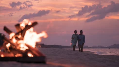 A couple walks along a beach at sunset, with a bonfire in the foreground and colorful sky in the background.