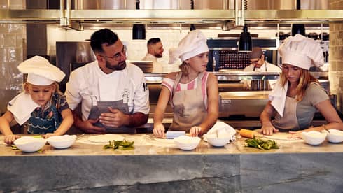 A chef standing at a counter teaching children wearing aprons and chef hats how to roll out pizza dough using rolling pins.