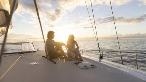 Couple in silhouette sitting barefoot on a boat deck framed by rigging ropes against a bright blue clouded sky at sunset.