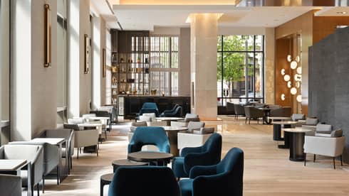 A large dining area with blue and light grey seats with round or square tables around them, large windows are letting in sunlight.