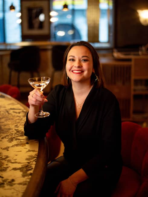 Camille Siralut in a black dress, sitting and smiling while holding a glass of champagne