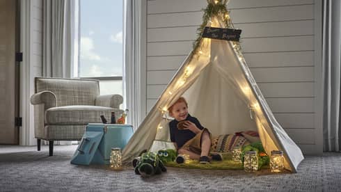 In a small tepee, a smiling child holds a cookie and is surrounded by pillows, stuffed animals and jars of twinkling lights.