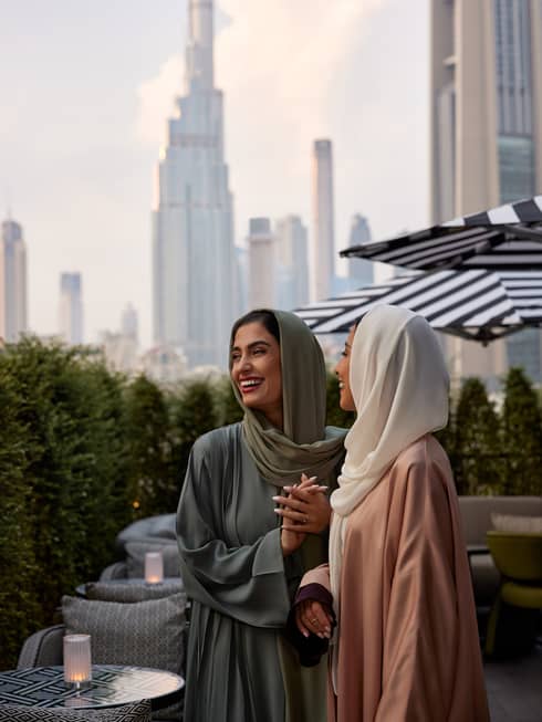 A woman wearing a sage-green hijab and dress stands close to another woman wearing a white hijab and pink dress on an outdoor terrace with the Dubai skyline in the background