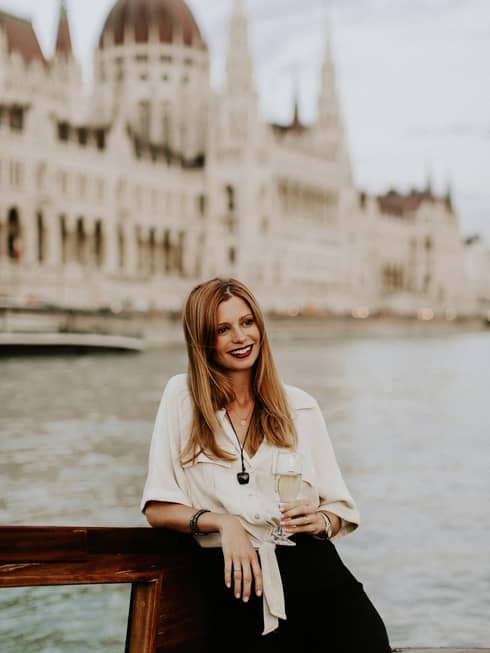 Long-haired women wearing ivory blouse and with glass of champagne in hand leaning on railing overlooking the Danube and Parliament building