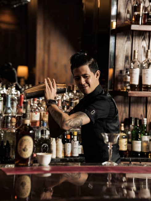 Bartender with short black hair wearing a black button-down short-sleeve shirt stand behind the bar shaking a silver cocktail shaker