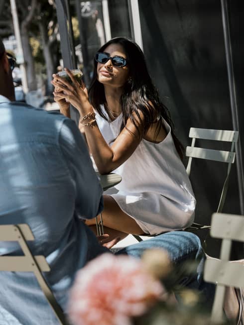 Person with long, straight dark hair wearing a white sleeveless mini dress and sunglasses sits at a bistro table by a sidewalk cafe