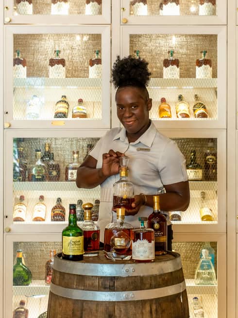 A woman in a grey shirt holding a bottle of liquor with shelves of liquor behind her.