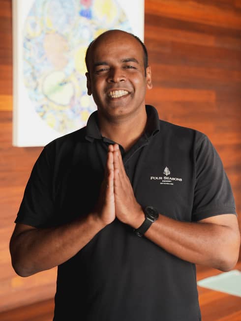 Justin James in black polo shirt, standing, smiling with namaste hands with wood-paneled wall and artwork in backdrop
