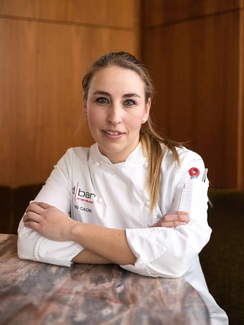 A woman with blonde hair wearing a chef's uniform stands in front of a brown wall.