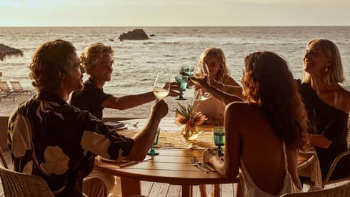 Five guests raise their glasses in a toast around a round wood table on a wood deck overlooking the beach and ocean.