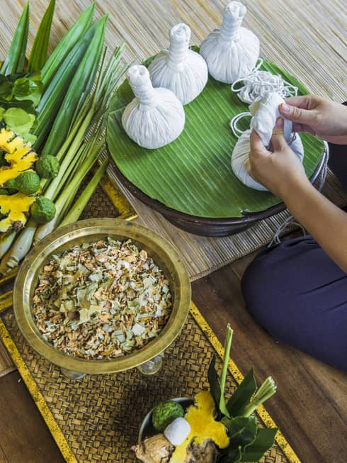 View over bowl of herbs, dried flowers as hands wrap cloth herbal pouches by spa tray