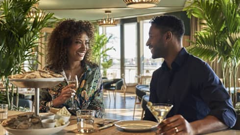 A man and woman eating oysters at an oyster bar with plants behind them.