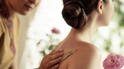 Spa staff rubs thick lotion on woman's bare shoulders as she holds a fresh flower