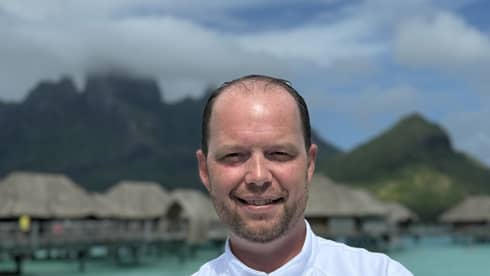 Smiling chef in white coat stands with arms crossed against backdrop of overwater pavilions