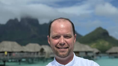 Smiling chef in white coat stands with arms crossed against backdrop of overwater pavilions