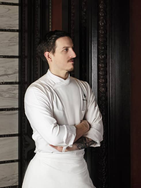 Chef de Cuisine Matteo Re Depaolini in white chef?s coat leans against tall wooden door with arms crossed