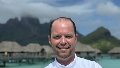 Smiling chef in white coat stands with arms crossed against backdrop of overwater pavilions