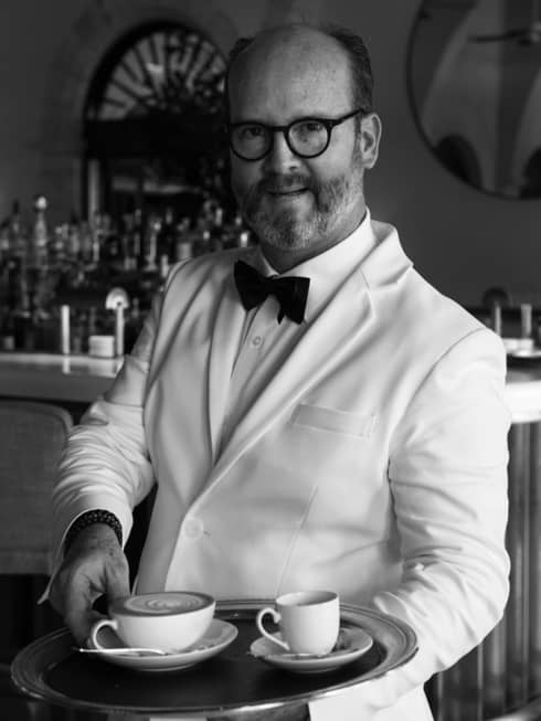 Man in white jacket and bow tie serves cappuccino at luxury hotel bar