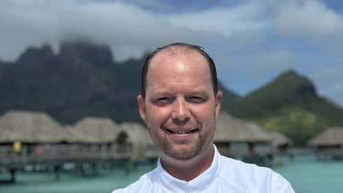Smiling chef in white coat stands with arms crossed against backdrop of overwater pavilions