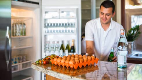 Smiling man holds green bottle in front of open fridge, fresh eggs on counter