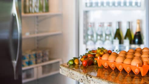 Smiling man holds green bottle in front of open fridge, fresh eggs on counter