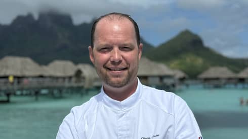 Smiling chef in white coat stands with arms crossed against backdrop of overwater pavilions