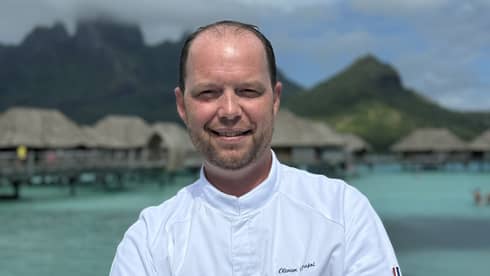 Smiling chef in white coat stands with arms crossed against backdrop of overwater pavilions