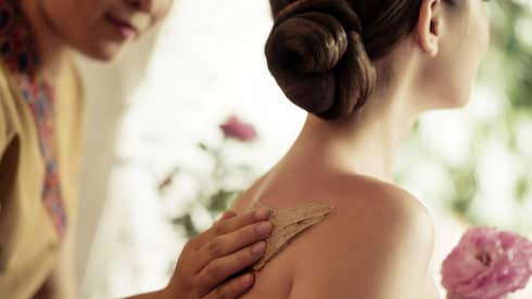 Spa staff rubs thick lotion on woman's bare shoulders as she holds a fresh flower