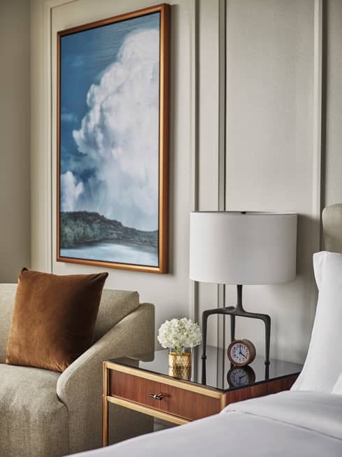 Framed painting of clouds over lake above armchair, nightstand with lamp, clock