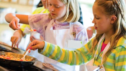 Two young girls in aprons stir pan on stove during Thai cooking class