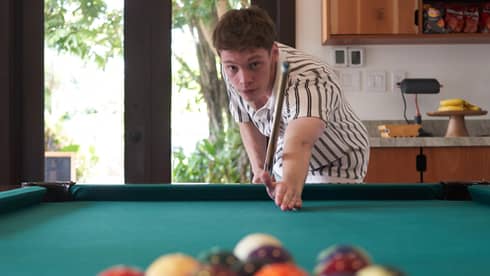 Teen wearing green-and-white button down short-sleeve shirt aims a cue at a collection of billiard balls on a green pool table