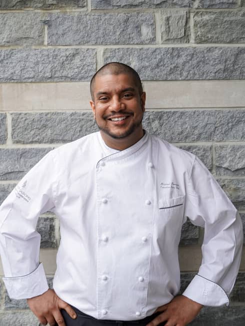 A man in a white chef's coat standing in front of a grey brick wall.