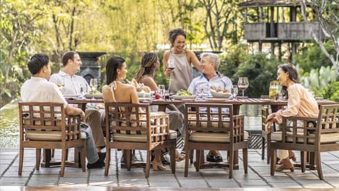 Group sharing a meal and drinks on an outdoor terrace with a chef grilling nearby, surrounded by lush greenery.