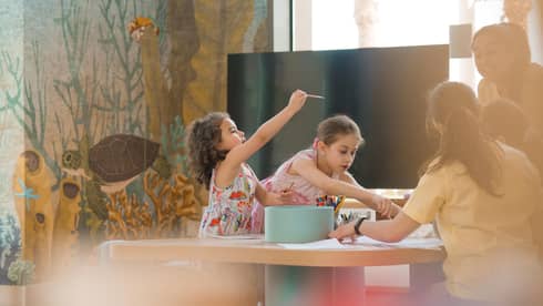 Two young children sit at a crafts table while an adult helps them paint