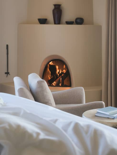 Hotel bed pillows, white armchair in front of curved clay fireplace, vases on mantle