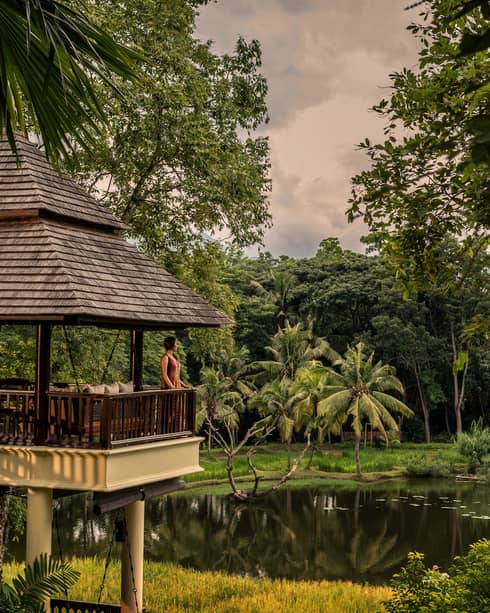 Person standing on a pavilion balcony overlooking a pond and tropical forest at sunset.