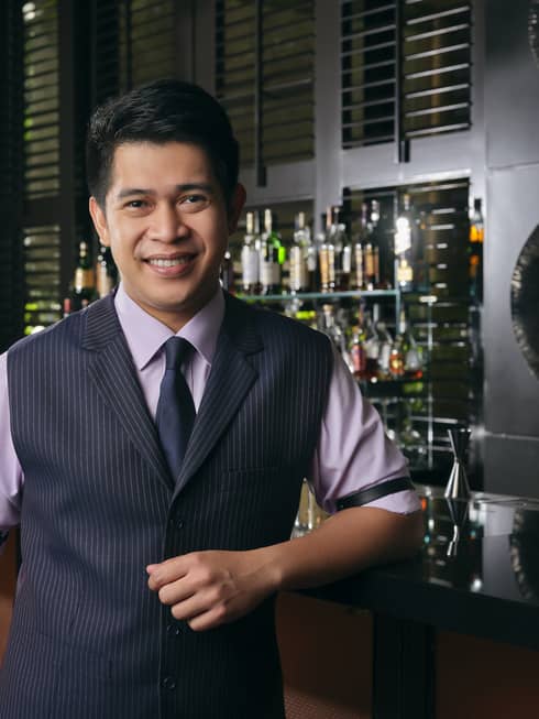 Smiling Bar Manager Gabriel Carlos poses for the camera, an artful display of liquor bottles on the shelf behind him