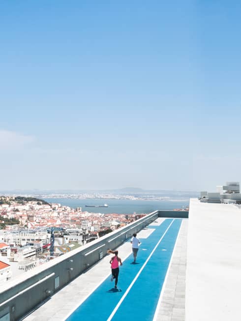 Aerial view of backs of man and woman running on blue rooftop outdoor track, Lisbon city roofs below