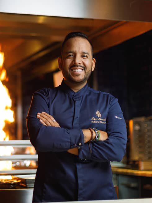 Chef wearing navy blue long-sleeve, button-up shirt stands with arms crossed in front of a kitchen with a large open flame in the background