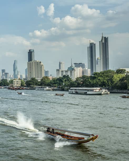 Longtail boat heading into foreground away from Bangkok skyline, leaving trail behind and surrounded by tall, splashing wake.