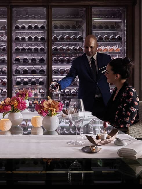 Man scans glass cellar as waitstaff pours wine in glass for woman at table