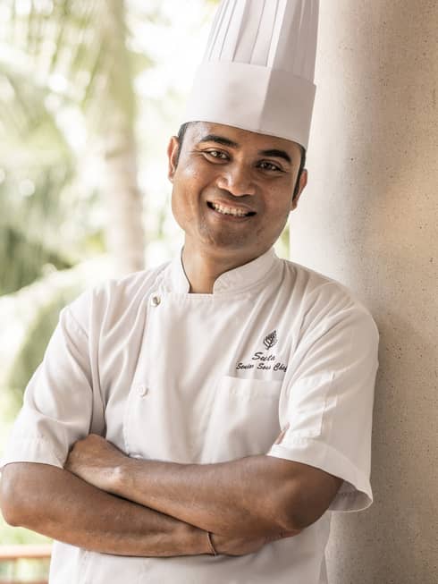 Smiling person in white chef's jacket and hat stands with arms crossed on balcony