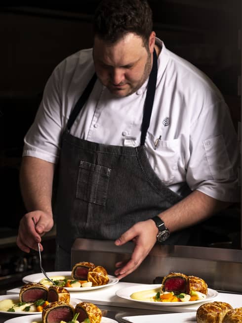 Two chefs in white shirts and blue aprons work on preparing several dishes of food.