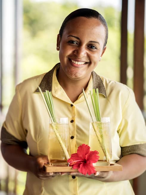 Smiling residence villa host in uniform holds tray with two cocktails, red tropical flower