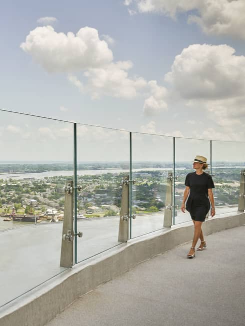A woman in black clothes walking along a glass wall of an observation deck looking out at a river.
