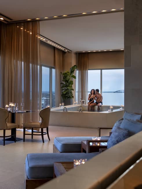 A couple enjoying a candlelit hot tub in a luxury hotel spa, with elegant seating, soft lighting and large windows offering a view of a bridge in the distance