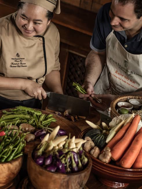 A chef teaching two participants in a cooking class, surrounded by fresh vegetables and ingredients in a rustic kitchen setting.