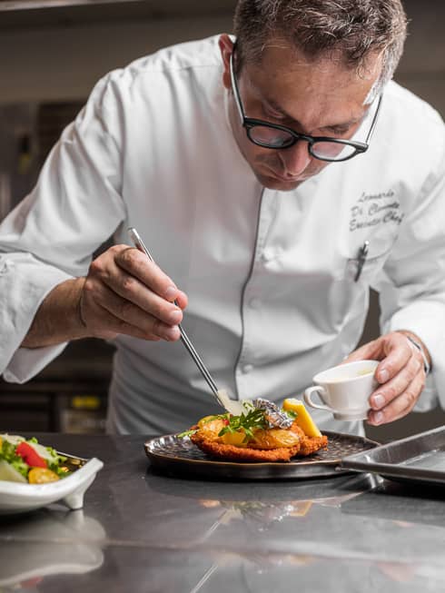 A chef lightly pours a sauce onto a plate of chicken and herbs.