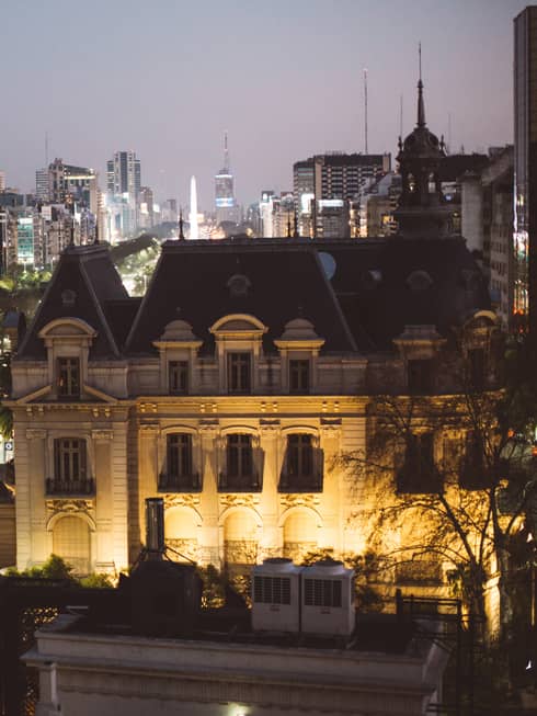 Exterior of Four Seasons Hotel Buenos Aires lit up at night next to a tree-line avenue and various city buildings across the street